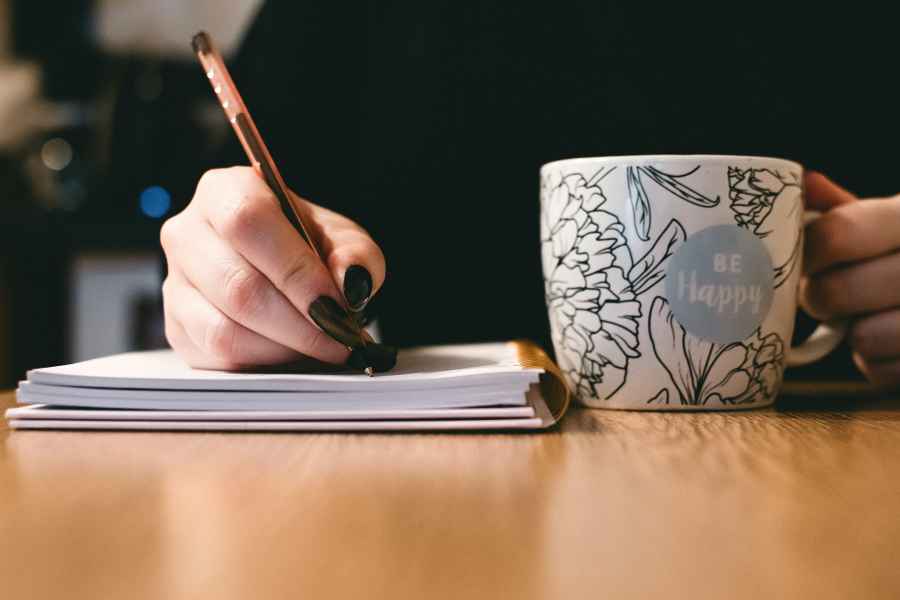 hand with black nail polish writing in notebook with a mug